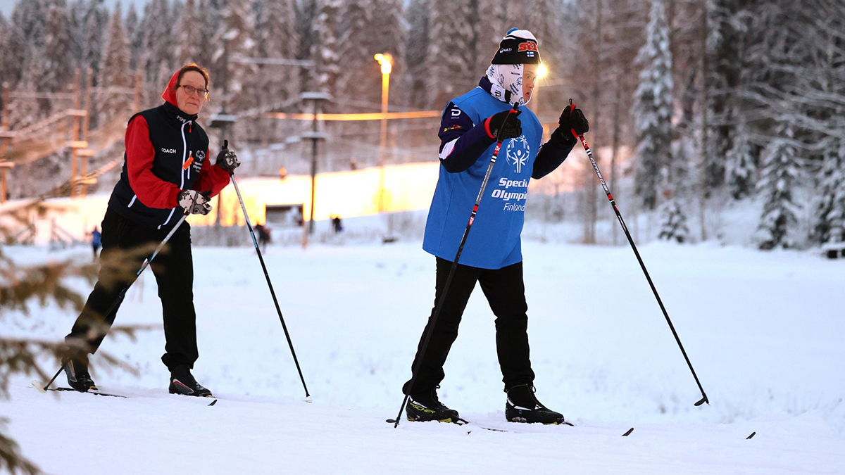 Mika Nyberg eteenpäin keskittyneenä ja Eeva Nyberg vähän matkan päässä perässä hiihtämässä auringonlaskussa. Kuvan molemmilla reunoilla näkyy lumisia havupuun oksia.